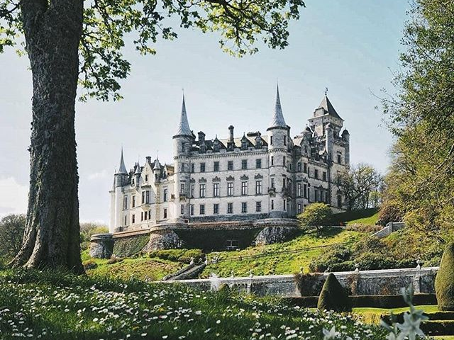 Em um castelo mesmo, com esculturas detalhadas, grandes jardins e no topo de uma colina ao lado do mar.