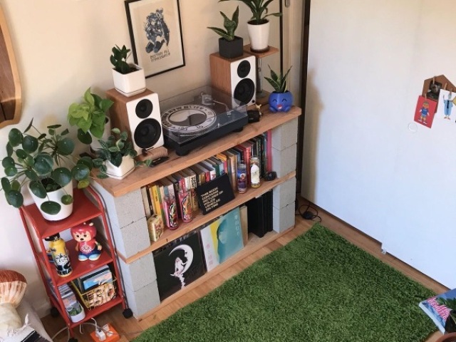 ♡ um quarto cheio de plantas, com outros tons verdes espalhadas pelo quarto. Rádios e uma vitrola, todos em cima de uma mesa, mais uns 100 livros. A sua cama e do lado da sua janelona, que também é cheio de plantas.