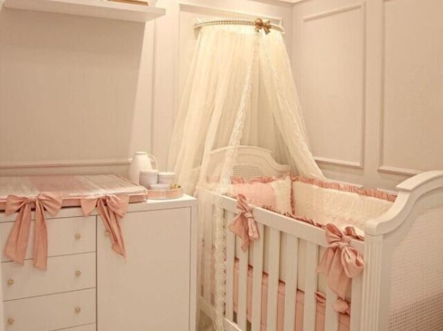 Quarto médio de tons brancos e rosa, berço com cortina .